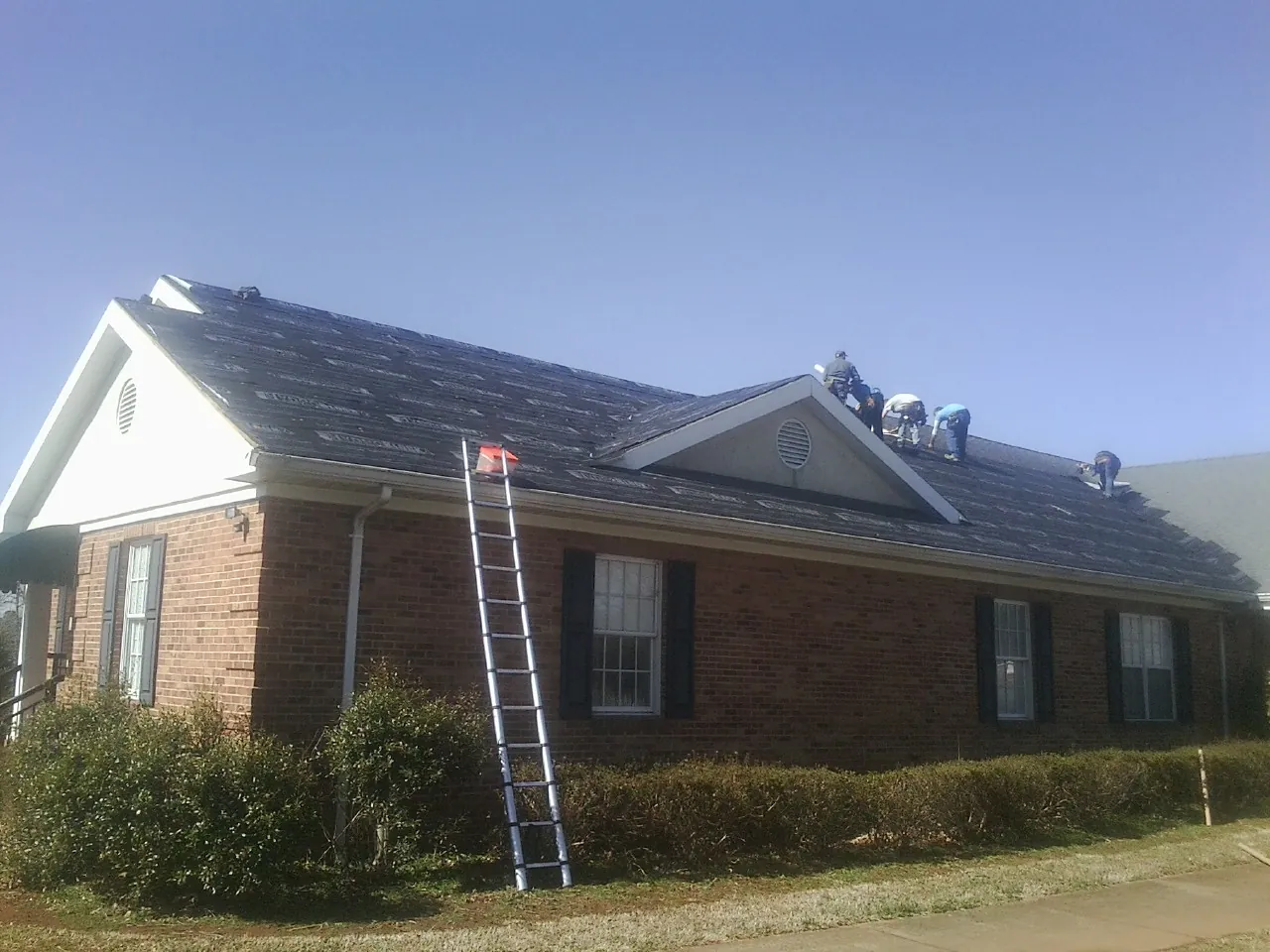 Crew tearing shingles off a brick ranch roof with a ladder set — Emergency Roof Repair work by our crew in East Kingston