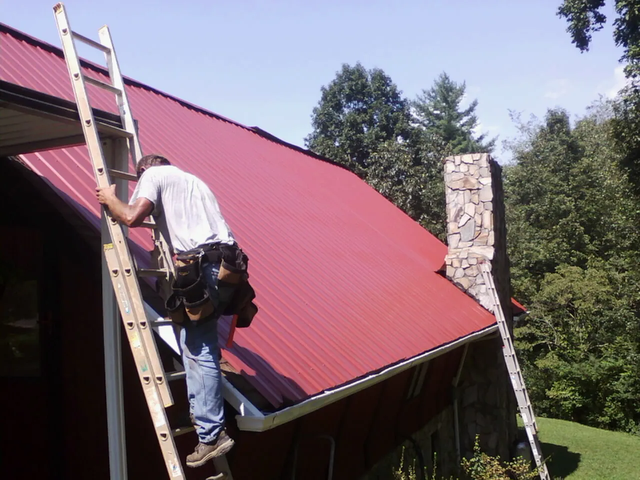 Roofer on a ladder beside a red metal roof and stone chimney — Soffit & Fascia Repair by Roofing Brothers in East Kingston